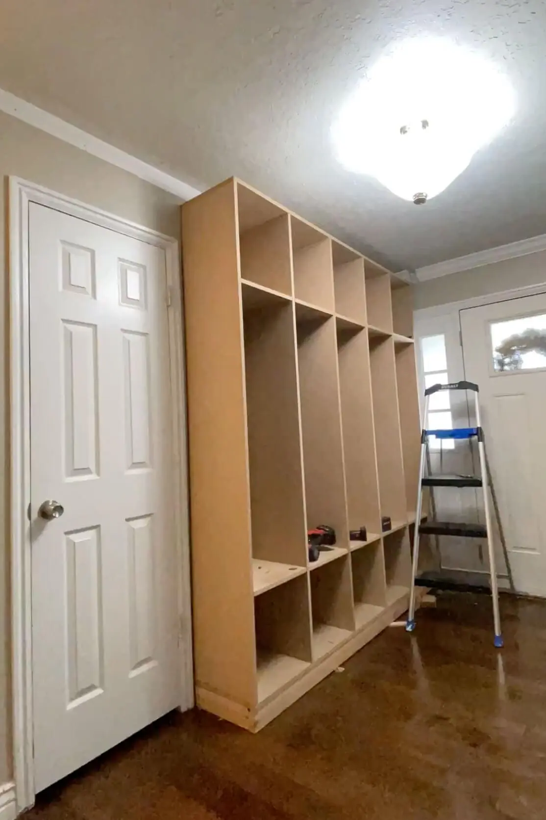 Partially completed DIY mudroom lockers installed in an entryway, featuring open cubbies and overhead storage, with a ladder nearby for final adjustments.